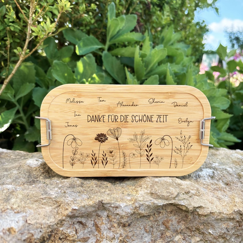 May include: A rectangular bamboo lunch box with a floral design and the text "DANKE F&Uuml;R DIE SCH&Ouml;NE ZEIT". The lid features names and metal clasps. The box is resting on a stone surface.