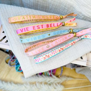 Könnte beinhalten: Fünf bunte Armbänder mit goldenen Buchstaben und Perlen. Die Armbänder sagen "Faith Hope", "Vie Est Belle", "Vires Gud", "Live Laugh" und "La".