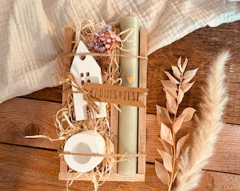 May include: A wooden box filled with a white ceramic house, a green candle, and dried flowers. The box has a tag that says "Frohes Fest" which is German for "Happy Holidays".