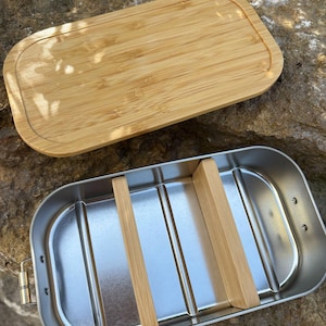 May include: A rectangular stainless steel lunch box with a bamboo lid. The interior has two bamboo dividers. The lid is made of light brown bamboo. The lunch box is open, revealing the interior.