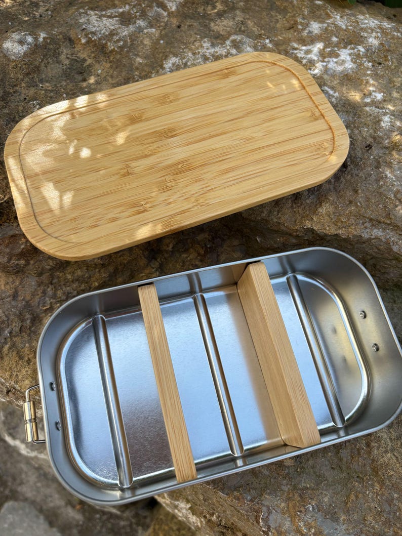May include: A rectangular stainless steel lunch box with a bamboo lid. The interior has two bamboo dividers. The lid is made of light brown bamboo. The lunch box is open, revealing the interior.