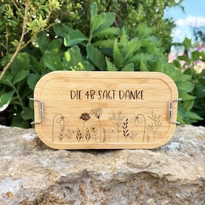 May include: A rectangular bamboo lunch box with rounded edges. The lid features the text "DIE 4B SAGT DANKE" and a floral design. The box has metal clasps on each side. The background is a natural setting with greenery.
