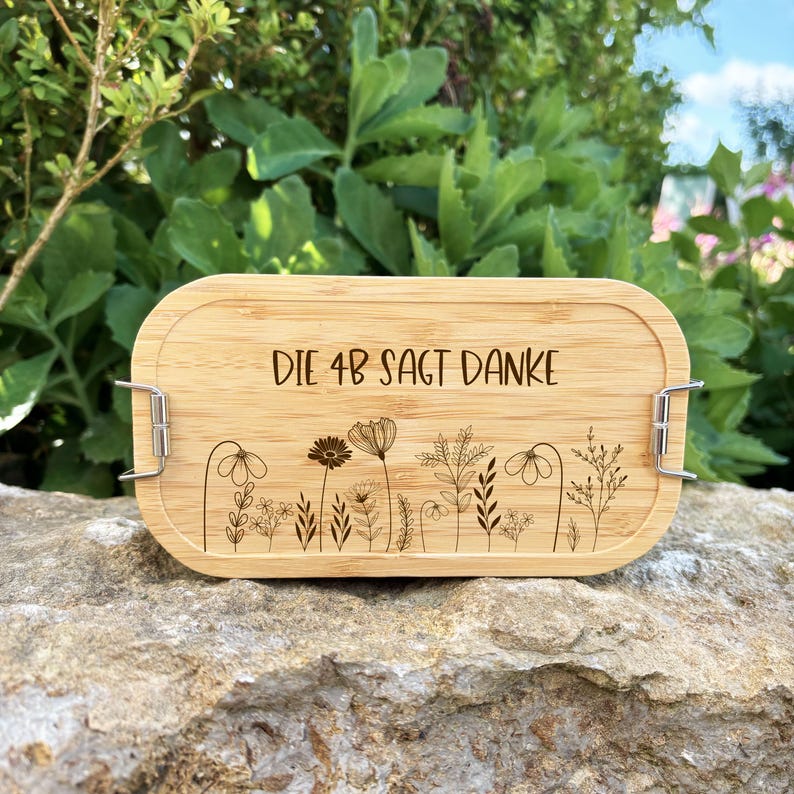 May include: A rectangular bamboo lunch box with rounded edges. The lid features the text "DIE 4B SAGT DANKE" and a floral design. The box has metal clasps on each side. The background is a natural setting with greenery.