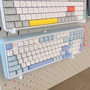 May include: A mechanical computer keyboard with a light blue frame and white keycaps featuring light blue accents on the modifier keys, including the escape, shift, enter, and arrow keys, mounted on a white pegboard wall display system.