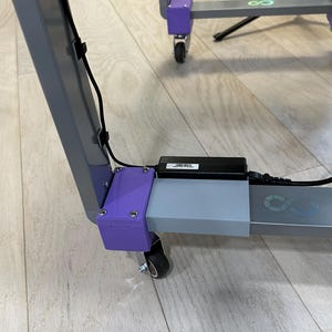 May include: A gray metal frame with purple corner supports and black wheels. A black power supply is attached, with a label. The frame has a logo and is on a light wood floor.