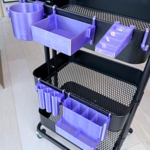 May include: A black rolling cart with various purple 3D-printed organizer attachments. The attachments include a cup holder, a rectangular tray, a tiered shelf, and several pen holders. The cart has a wire mesh design on the shelves.