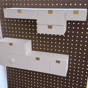 May include: A collection of white storage drawers with gold-colored recessed handles, mounted on a brown pegboard. The drawers are arranged in various configurations, offering a modular storage solution. The pegboard has evenly spaced holes.