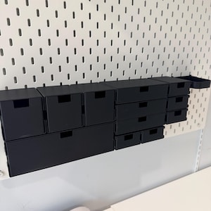 May include: A collection of black storage boxes and drawers of various sizes mounted on a white pegboard. The drawers have a minimalist design with a small rectangular opening for easy access. The pegboard is attached to a white wall.