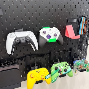 May include: A black pegboard displaying several video game controllers. Controllers include a white and black PlayStation 5 controller, a white and black Xbox controller, and several colorful controllers. A pink phone stand and cable organizers are also visible.