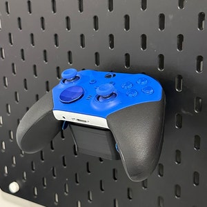 May include: A blue Xbox controller with black grips and buttons is mounted on a black pegboard wall.