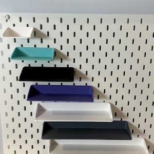 May include: A white pegboard with vertical slots featuring seven attached shelves in various colors including white, turquoise, black, purple, and dark gray, arranged in a descending diagonal pattern across the surface of the board.