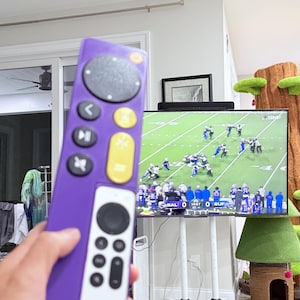 May include: A purple remote control with a circular top button and several other buttons in yellow and white. The remote is in front of a television screen displaying a football game.