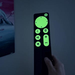 May include: A black remote control with glowing green buttons and symbols. The remote has a power button, volume controls, and playback buttons. The remote is held in a hand against a dark background.
