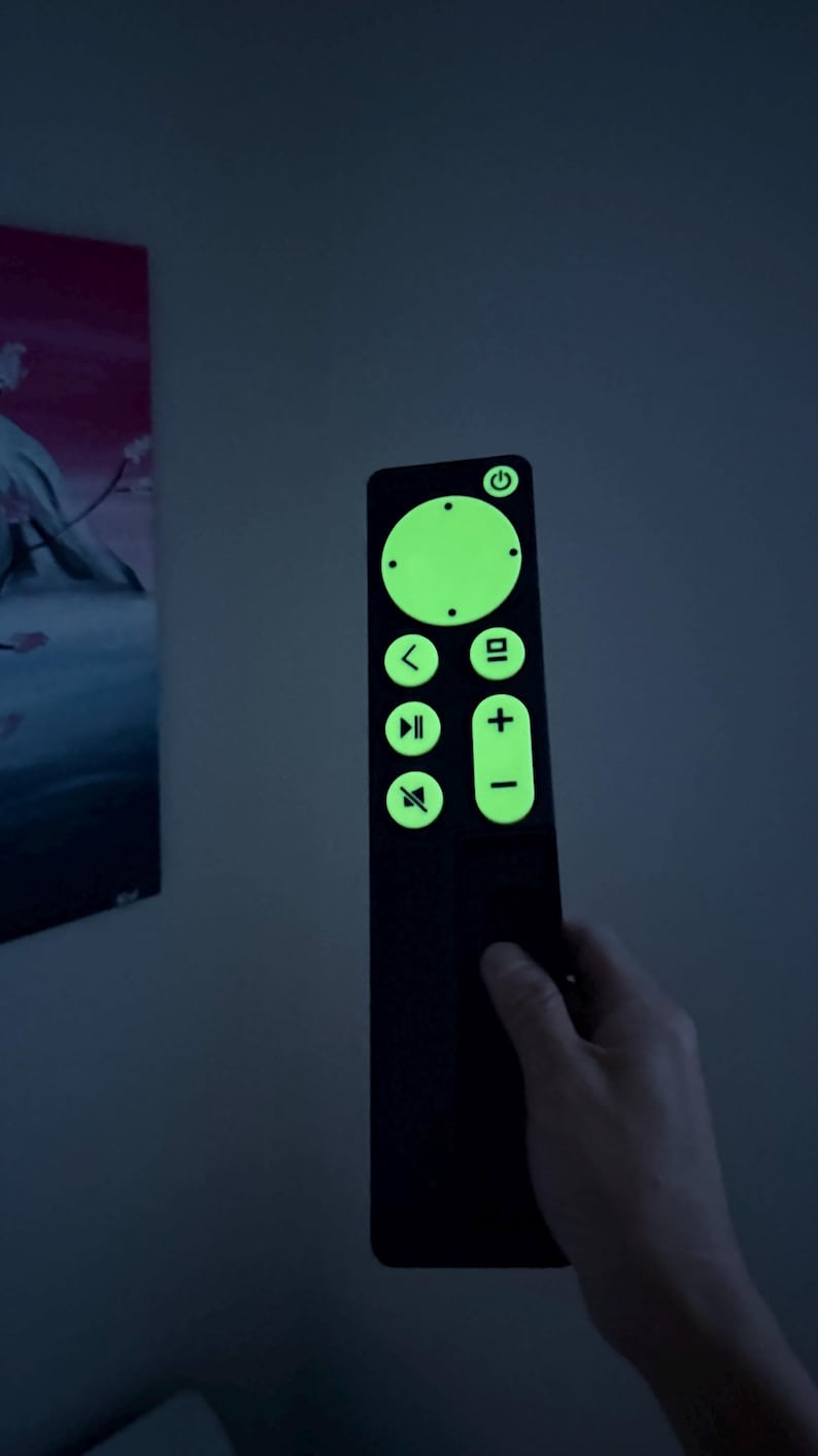 May include: A black remote control with glowing green buttons and symbols. The remote has a power button, volume controls, and playback buttons. The remote is held in a hand against a dark background.