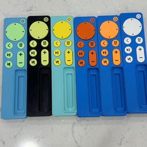 May include: A collection of six colorful remote control covers. The covers are in various colors, including light blue, black, teal, yellow, orange, and white. Each cover has a circular button and several directional buttons.