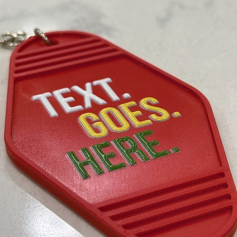Puede incluir: Un llavero estilo motel de color rojo con superficie texturizada y anilla met&aacute;lica. El llavero presenta el texto en relieve blanco "TEXT." seguido del texto en relieve amarillo "GOES." y el texto en relieve verde "HERE." en la parte frontal.