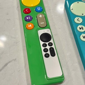 May include: Two colorful TV remotes. One is green with a yellow circle and multi-colored buttons. The other is teal with a light green circle and white buttons. Both have a white section with black buttons.