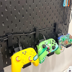 May include: A black pegboard wall organizer holding three video game controllers. One controller is bright yellow with a smiling face, one is green with a Yoshi character graphic, and one is a clear plastic shell with visible internal components.
