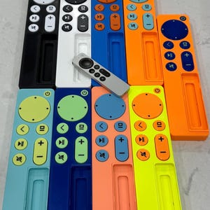 May include: A collection of colorful remote control cases in various shades, including black, white, blue, orange, and green. Each case has a rectangular design with button cutouts and a central slot. A silver remote is also present.