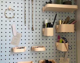 May include: A light blue pegboard wall organizer featuring cream-colored modular shelves, hooks, and bins. The system holds various items including necklaces, hoop earrings, makeup brushes, lipsticks, and small jewelry pieces, with two paper notes attached.