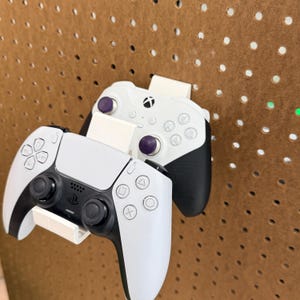 May include: Two white and black game controllers, one with purple accents, are mounted on a brown pegboard. The controllers are for different gaming consoles. The controllers are held in place by white brackets.