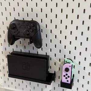 May include: A black Nintendo Switch console and two controllers are mounted on a white pegboard wall. The console is in a black stand and the controllers are in a black stand with a mint green and pink controller.
