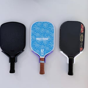 May include: Three pickleball paddles mounted on a wall. The left is solid black. The center paddle features a blue pattern with smiley faces and the text Bread & Butter Pickleball Co. The right paddle is black with Joola branding and red accents.