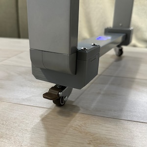 May include: A close-up of a gray, metal, adjustable desk frame with wheels. The frame has a rectangular shape with a matte finish. The wheels are small and brown. The desk frame is on a light-colored wooden floor.