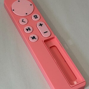 May include: A pink remote control with a power button, directional pad, and volume controls. The remote has a rectangular shape with rounded edges and a recessed area at the bottom. The buttons are black and the remote is made of plastic.
