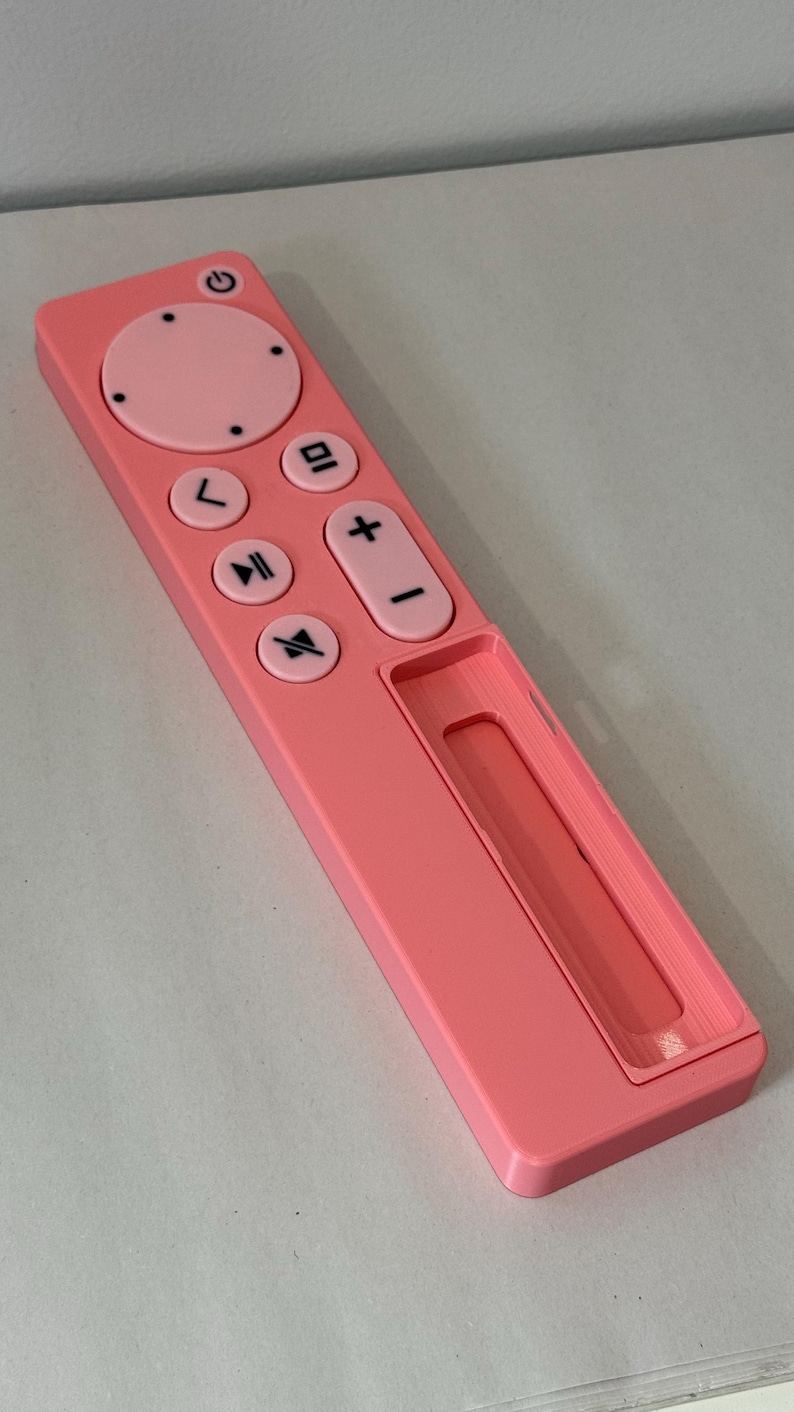 May include: A pink remote control with a power button, directional pad, and volume controls. The remote has a rectangular shape with rounded edges and a recessed area at the bottom. The buttons are black and the remote is made of plastic.