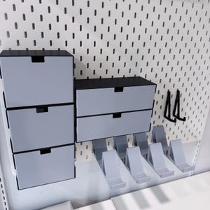 May include: A modular storage system mounted on a white pegboard, featuring three stacked black and light gray drawers on the left, two side-by-side drawers on the right, four light gray angled shelf brackets, and two black L-shaped hooks.