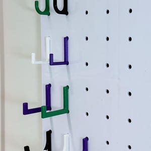 May include: Four sets of hooks in different colors, including black, white, green, and purple, are mounted on a white pegboard. The hooks are all in the shape of a 'J' and are arranged in rows.