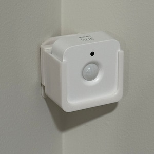 May include: A white Philips Hue motion sensor mounted on a white wall. The sensor has a round, white motion detection lens.