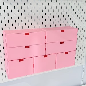 May include: A set of eight pink storage boxes with red pull tabs, mounted on a white pegboard. The boxes are rectangular and stacked in a grid pattern, offering a clean and organized look for storage.