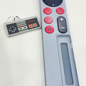 May include: A light gray remote control with a black circular power button and red circular buttons. A small Nintendo controller is next to the remote. The remote has a plus and minus button and a rectangular opening.