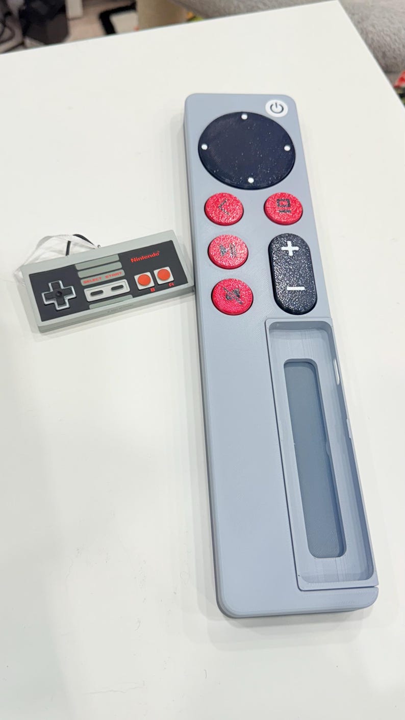 May include: A light gray remote control with a black circular power button and red circular buttons. A small Nintendo controller is next to the remote. The remote has a plus and minus button and a rectangular opening.