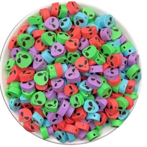 Puede incluir: Un bol lleno de cuentas de plástico con forma de cara de extraterrestre de colores. Las cuentas son de plástico y vienen en verde, azul, rojo y morado.