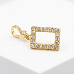 May include: A gold-colored rectangular pendant with a halo of small, clear gemstones. The pendant is connected to a small, circular loop with a single gemstone. The jewelry is displayed on a white surface.