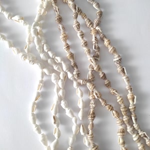 Könnte beinhalten: Ein Set aus fünf Strängen weißer und brauner Muschelketten. Die Ketten bestehen aus kleinen, gleichmäßigen Muscheln, die auf einem durchsichtigen Faden aufgereiht sind.