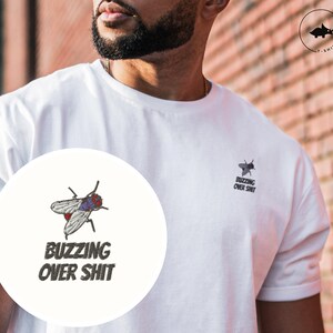 May include: White t-shirt with a black and gray fly graphic and the text "Buzzing Over Shit" on the front. The shirt is being worn by a person with a beard.