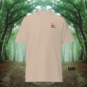 Puede incluir: Una camiseta beige con un peque&ntilde;o gr&aacute;fico de zorro bordado y el texto "For the Same" en la parte delantera. La camiseta est&aacute; fotografiada sobre un fondo borroso de un camino en el bosque.