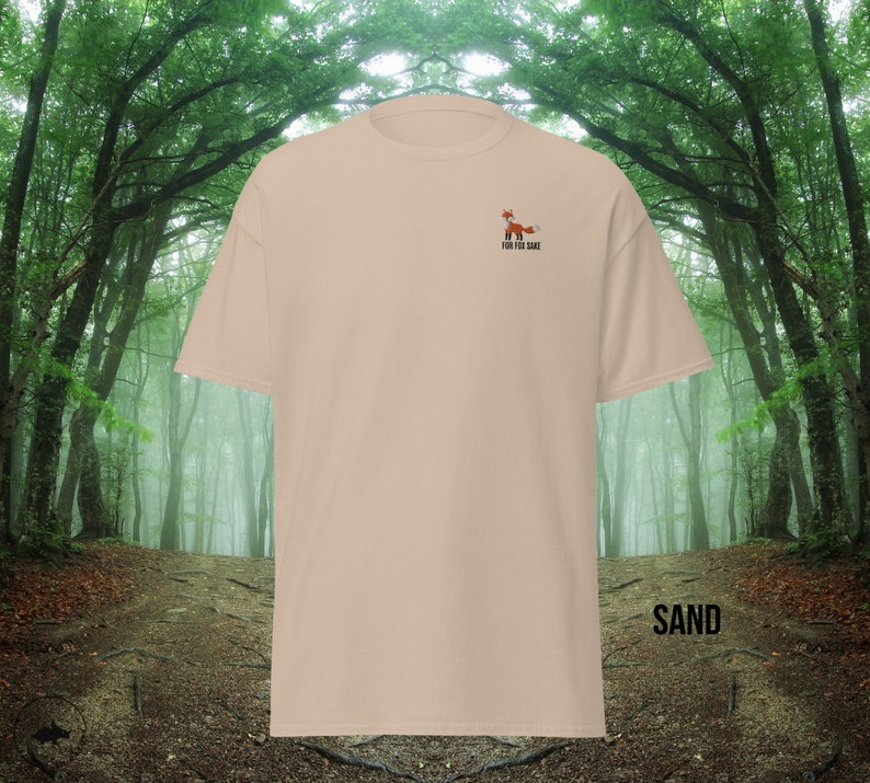 Puede incluir: Una camiseta beige con un peque&ntilde;o gr&aacute;fico de zorro bordado y el texto "For the Same" en la parte delantera. La camiseta est&aacute; fotografiada sobre un fondo borroso de un camino en el bosque.
