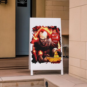 May include: A white sign with a red balloon and the text "You'll Float Too" on it. The sign features a scary clown with red hair and a red and white striped shirt. The clown is holding a yellow object in his hand.