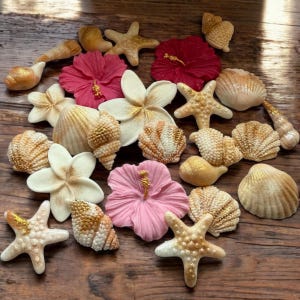 May include: A collection of decorative seashells, starfish, and flowers in different sizes and shapes. The shells and starfish have a gold glitter finish, while the flowers are pink, red, and white. They are arranged on a wooden surface.