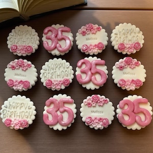 May include: Twelve round white cupcake toppers with scalloped edges. Some feature the silver text "happy BIRTHDAY" with pink flowers, others display the number "35" in metallic pink, and some are personalized with the name "Melissa" and pink flowers.