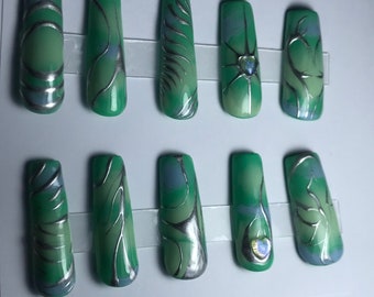 May include: Set of ten long, square-shaped artificial nails. Each nail is painted in shades of green with silver swirls and accents. Some nails feature a small, iridescent heart-shaped gem. The nails are arranged on a clear plastic strip.