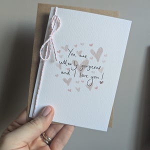 Könnte beinhalten: Eine Grußkarte mit der handgeschriebenen Botschaft "You are utterly gorgeous and I love you!", begleitet von Herzillustrationen. Die Karte ist mit rosa-weißem Garn gebunden.
