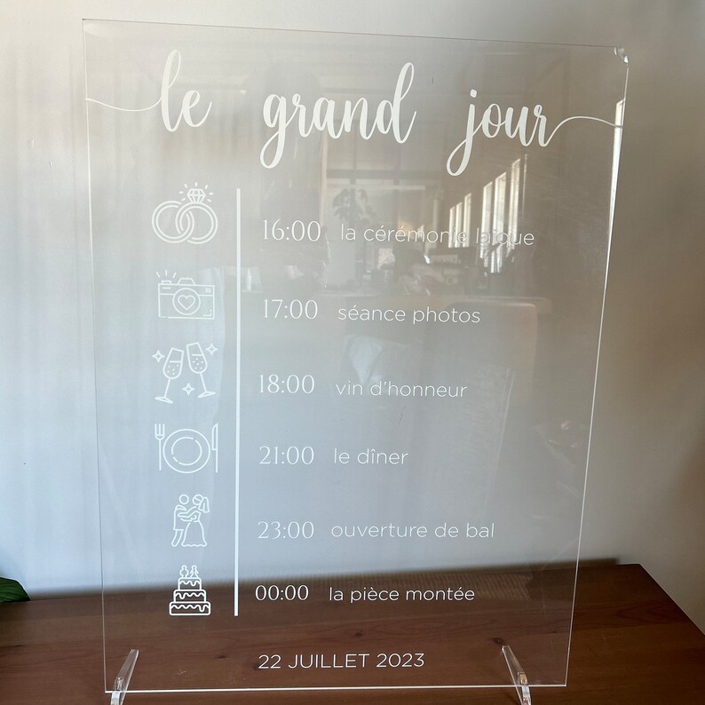 May include: A clear acrylic sign with a white inscription that reads "le grand jour" and lists the schedule for a wedding day. The schedule includes the ceremony, photos, dinner, and dancing. The date is July 22, 2023.