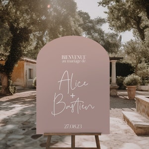 May include: A dusty rose arch-shaped welcome sign with white text that reads "Bienvenue au mariage de Alice + Bastien 27.08.23". The sign is on a wooden easel in front of a stone courtyard with a building and trees in the background.