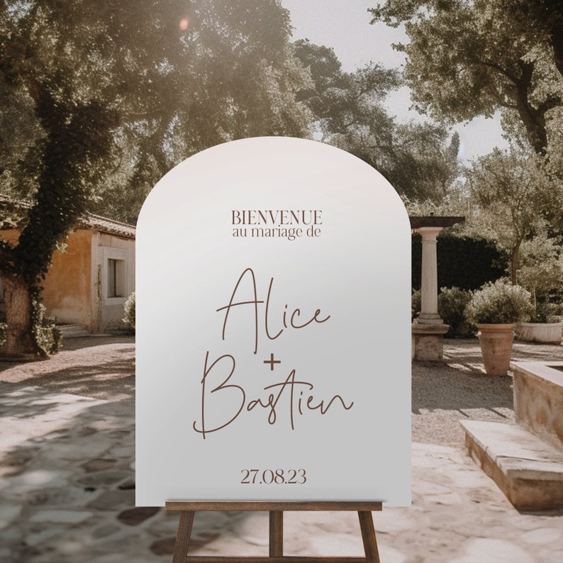 May include: White arch-shaped sign with brown text that reads "Bienvenue au mariage de Alice + Bastien 27.08.23". The sign is on a wooden easel in front of a stone building and lush greenery.